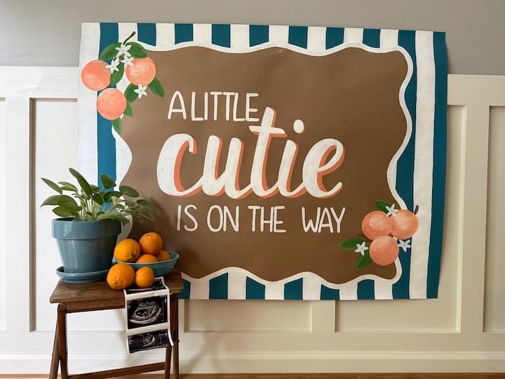 Hand-painted baby shower banner reading “A Little Cutie Is On The Way,” with citrus oranges, a blue stripe border, and ultrasound photos styled nearby.