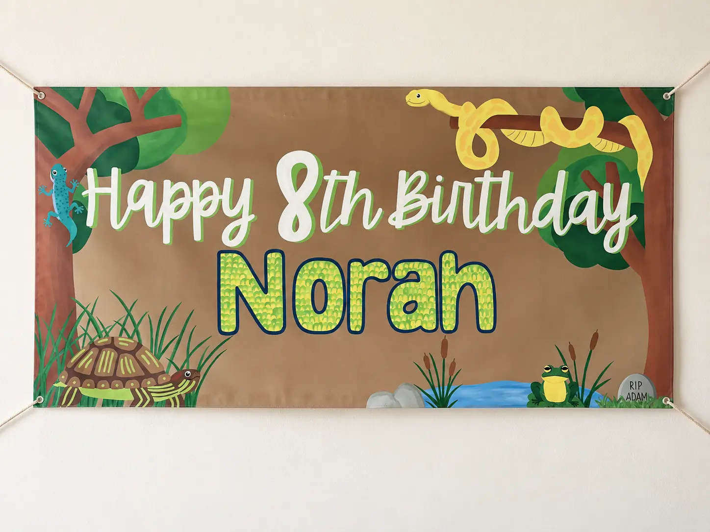 The “Happy 8th Birthday Norah” reptile-and-pond banner hanging on a wall against a cream background.