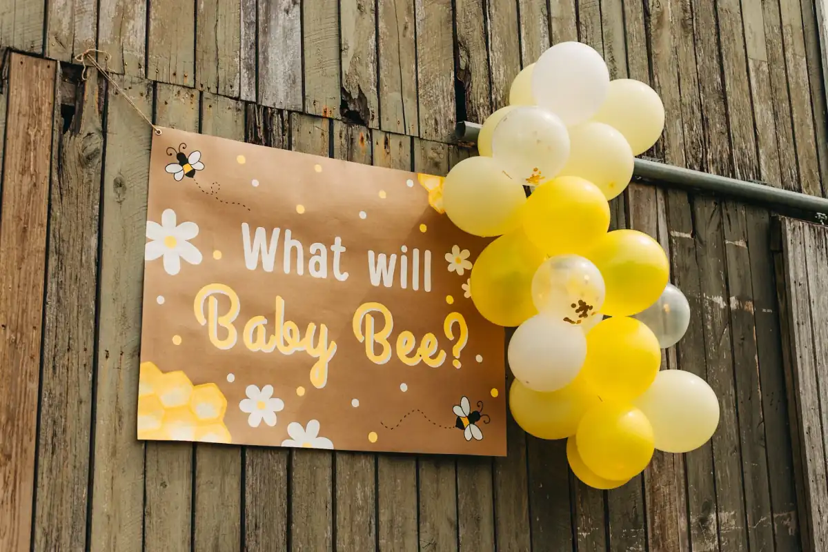 Hand-painted kraft banner reading “What will Baby Bee?” with bees, daisies, and honeycomb, hanging on a weathered barn next to a cluster of yellow and white balloons.