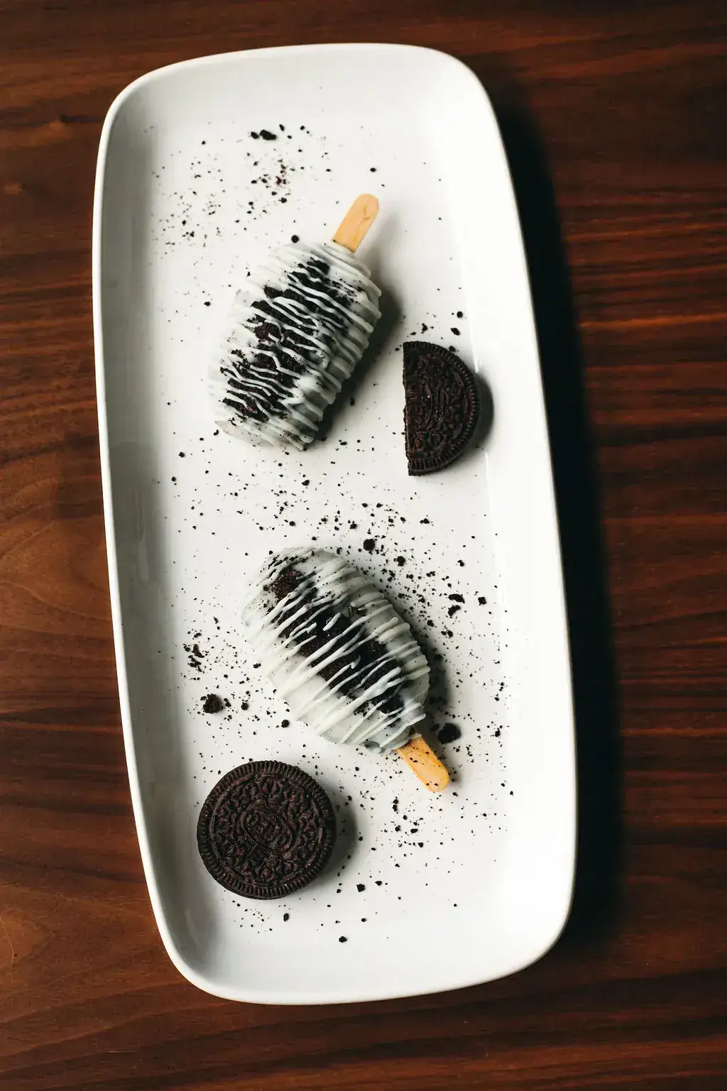 Two cookies-and-cream cakesicles on a rectangular white platter, styled with crushed chocolate sandwich cookies and cookie halves.