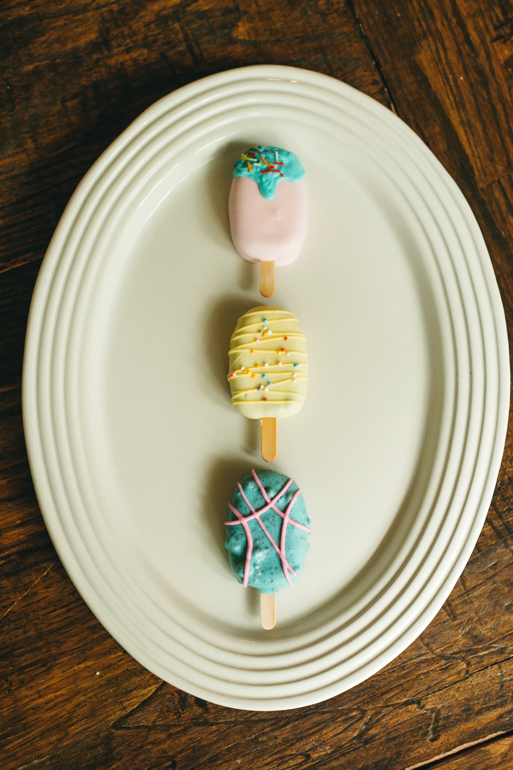 Three cakesicles on a white plate: a pink cakesicle with a teal drip and rainbow sprinkles, a pale yellow cakesicle with yellow drizzle and sprinkles, and a turquoise cakesicle with pink piped lines.