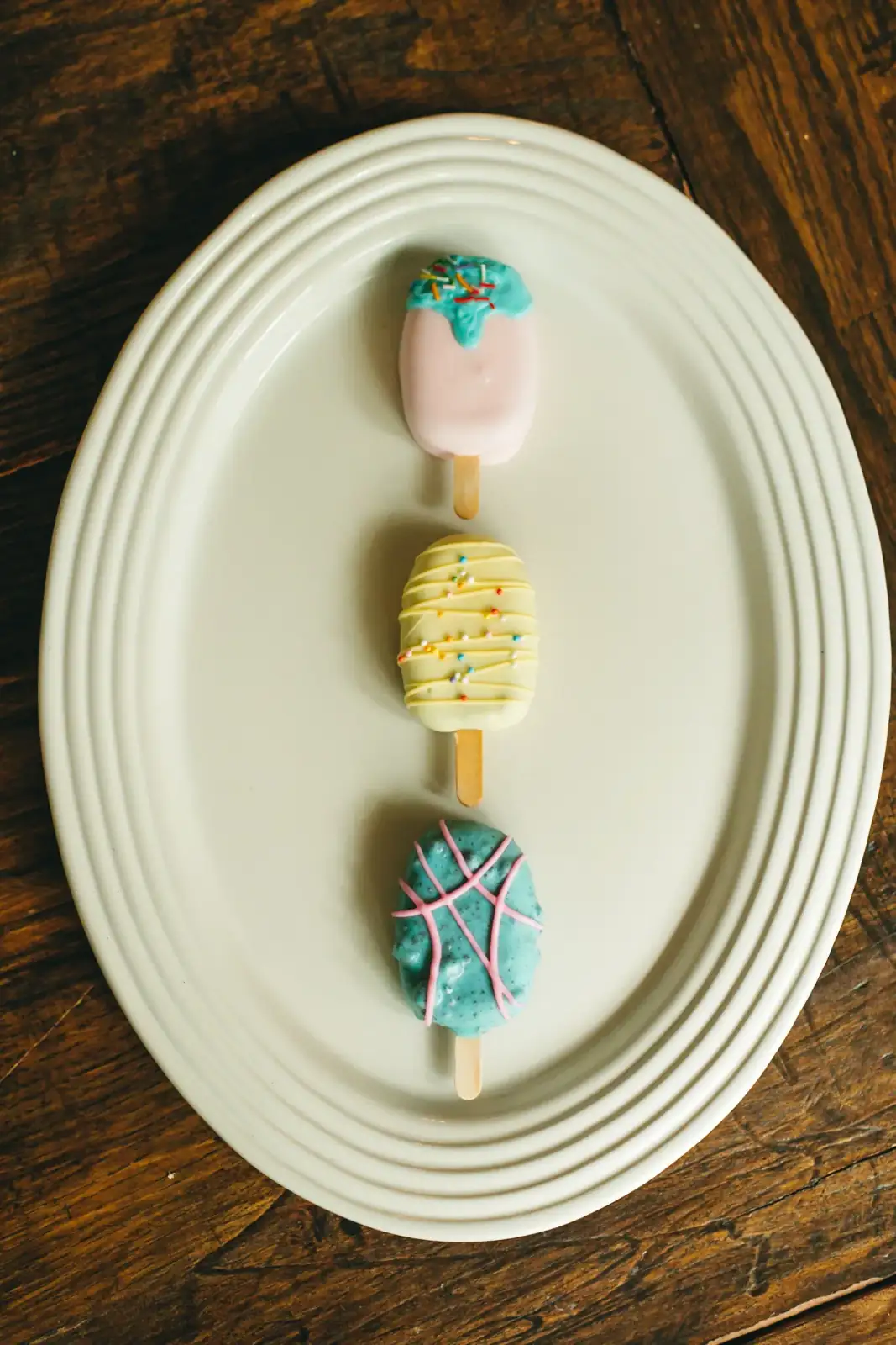 Three cakesicles on a white plate: a pink cakesicle with a teal drip and rainbow sprinkles, a pale yellow cakesicle with yellow drizzle and sprinkles, and a turquoise cakesicle with pink piped lines.