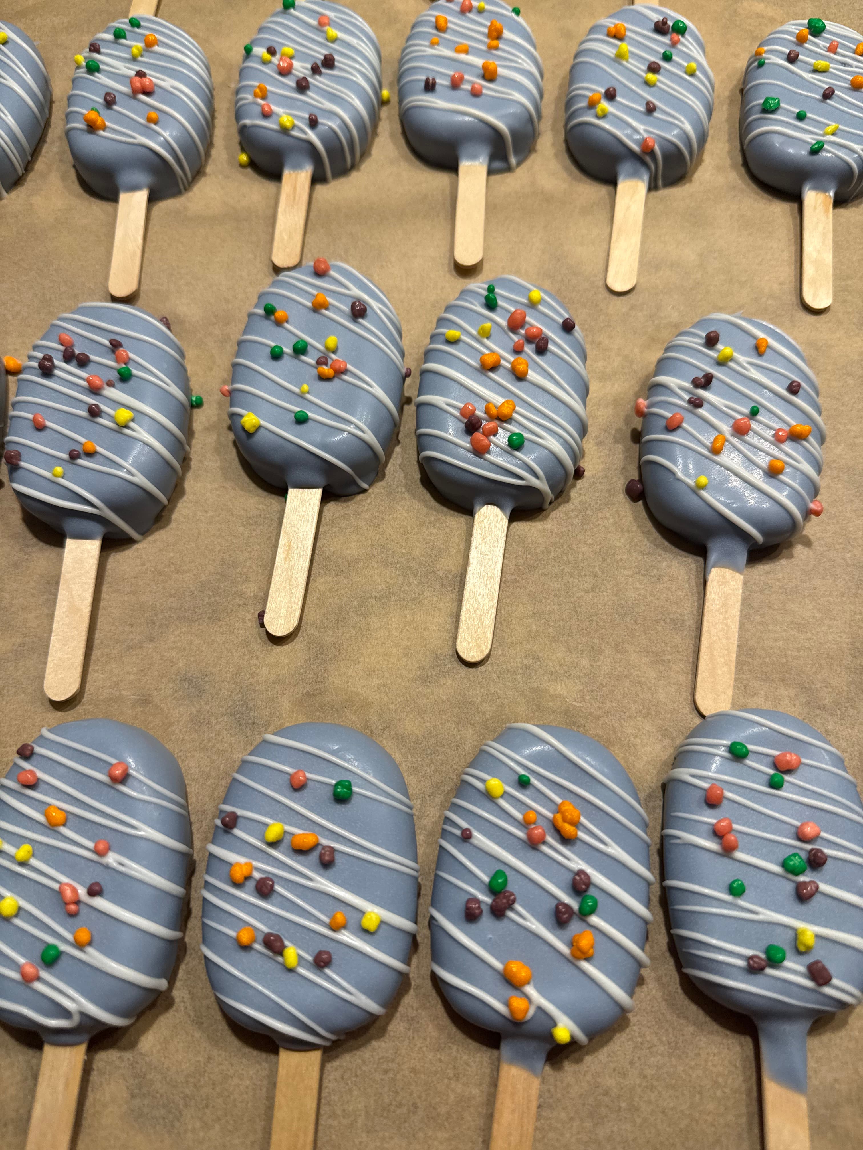 Overhead view of a batch of gray-blue cakesicles with white drizzle and colorful candy pieces, arranged in rows on parchment paper.
